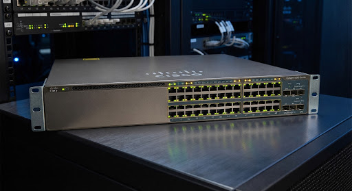 High-angle studio product shot of Cisco Catalyst 9300 48-port PoE enterprise network switch, metallic chassis front view with 48 RJ-45 copper ethernet ports glowing active green and amber LEDs, pristine condition on dark metallic surface with soft blue datacenter reflections